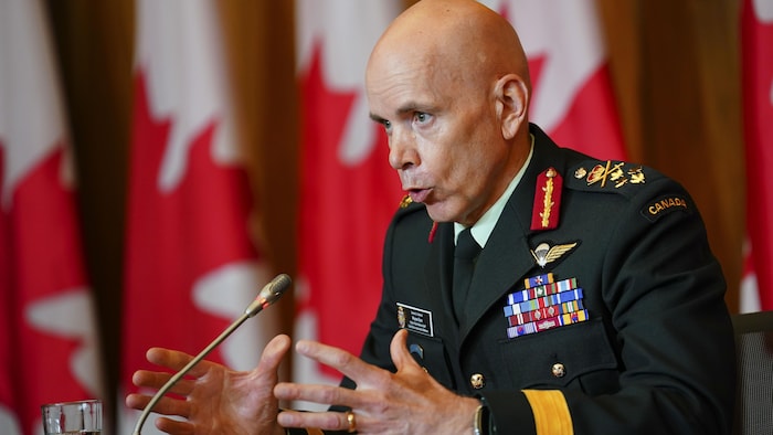 رئيس هيئة أركان الدفاع في القوات المسلحة الكندية، الجنرال واين إير، يتحدث في مؤتمر صحفي، أمامه ميكروفون وخلفه أعلام كندية. 
